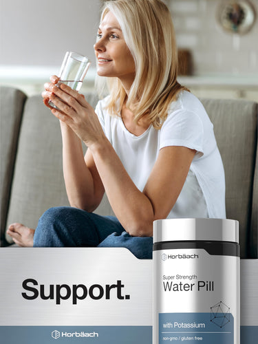 View 6 Middle-aged blonde woman smiles, sitting on a couch with a glass of water, next to a Horbaach Water Pill bottle.