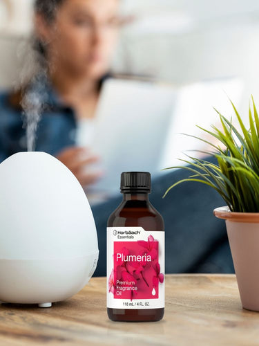 Bottle of Plumeria fragrance oil next to a diffuser on a table with a blurred person in the background.