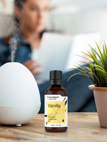 Bottle of vanilla fragrance oil next to a diffuser on a table with a blurred person in the background.