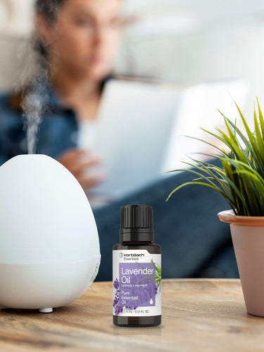 Bottle of lavender oil with a diffuser and plant on a table, person using laptop in background