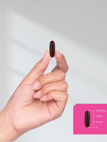 Hand holding a dark red pill with a size reference on a pink background.
