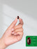 Hand holding a dark red capsule with size reference on a green background.