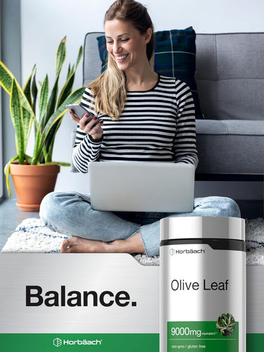 View 6 A young woman smiling while using her phone on the floor next to a laptop and an Olive Leaf bottle.